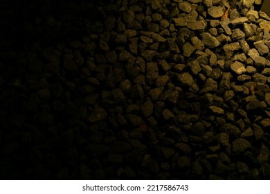 Small Rock Bed With Yellow Light Gradient. Batu Split Coffee Shop. Texture Of Ballast Gravel Stone On A Dark Black Background As Empty Negative Space. Tiny Rock Illuminated By Yellow Warm Light.