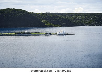 A small rocky island in the Oslo Fjord, Norway, features the Steilene Lighthouse and a white building, with a mountain in the background and the Fjellstrand village on the mountainside - Powered by Shutterstock