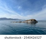 Small, rocky island surrounded by calm blue waters, with a mountainous background and soft clouds. A peaceful scene capturing the rugged beauty of a solitary island in a tranquil setting.