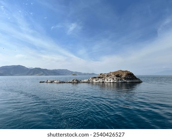 Small, rocky island surrounded by calm blue waters, with a mountainous background and soft clouds. A peaceful scene capturing the rugged beauty of a solitary island in a tranquil setting. - Powered by Shutterstock