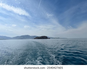 Small, rocky island surrounded by calm blue waters, with a mountainous background and soft clouds. A peaceful scene capturing the rugged beauty of a solitary island in a tranquil setting - Powered by Shutterstock