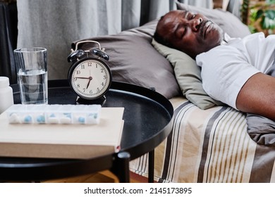 Small Round Black Table With Alarm Clock, Medicaments, Book And Glass Of Water Standing By Double Bed Where Senior Man Sleeping