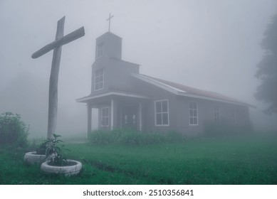 A small, rustic church surrounded by thick fog, with a simple wooden cross standing in front, creating a mysterious and serene atmosphere in a misty, rural setting. - Powered by Shutterstock