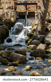 Small Rustic Waterfall Flowing Over Mossy Rocks Under Wooden Bridge Sluice Gate in Scenic Garden Park