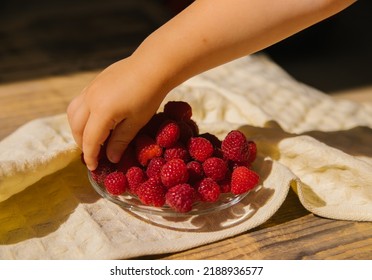 A small saucer with ripe raspberries, a bunch of summer berries on a wooden background. A child's hand takes a berry from the dishes. Summer background. Juicy appetizer or dessert.