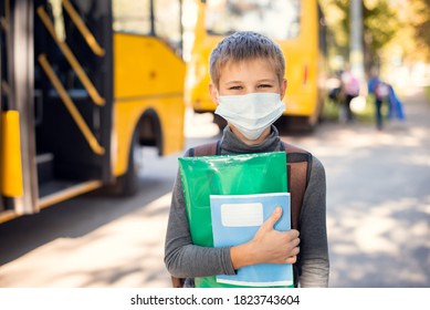 Kleiner Schuljunge in medizinischer Maske, der am Morgen in der Nähe eines Schulbusses Lernmaterialien hält. Konzept der Schulreise mit einem Schulbus während einer Pandemie