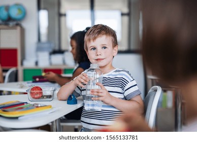 Small School Boy Sitting At The Desk In Classroom, Drinking Water.