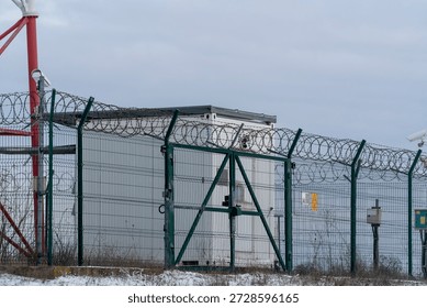 A small security building is surrounded by a tall fence with barbed wire. The setting is cold and wintery, and the sky is cloudy. The area appears isolated.