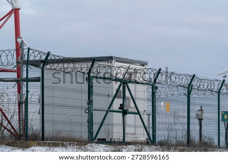 A small security building is surrounded by a tall fence with barbed wire. The setting is cold and wintery, and the sky is cloudy. The area appears isolated.