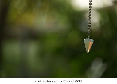 A small, shiny opalite pendulum with a silver chain is suspended against a soft, green background, evoking a sense of calm and spirituality.