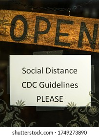 A Small Shop Displays An Open Sign Along With An Additional Request For Shoppers To Social Distance And Follow CDC Guidelines Due To The COVID-19 Pandemic.