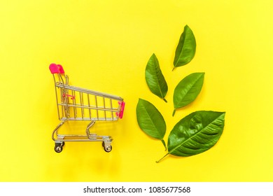 Small Shopping Trolley Cart Toy With Green Leaves On Yellow Colourful Trendy Modern Fashion Background. Ecology Eco Products Health Food Vegan Vegetarian Shopping Concept