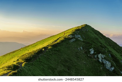 A small silhouette of a man in red walking along a mountain path on the slope of a huge mountain on a summer evening. The serene view of the green mountain against the sky is truly breathtaking - Powered by Shutterstock