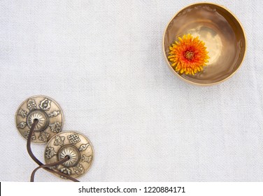 A small singing bowl stands in the upper right corner filled with water with floating chrysanthemum inside. A small singing bowl and tingsha stand on a gray background of organic cotton. Top view.