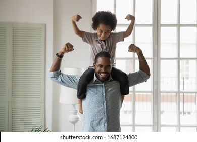 Small son sit on strong dad shoulders showing biceps. African family enjoy activity games at home, healthy fit lifestyle, two superheroes, vitamins for adults and children ad, happy Father Day concept - Powered by Shutterstock