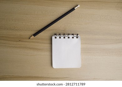 A small spiral notepad and a sharpened black pencil with an eraser lie on a wooden surface.