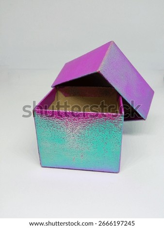 ​a small, square gift box, for a ring or other small piece of jewelry, placed on a plain white surface. The box is made of cardboard and is covered in an iridescent material that shines.