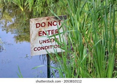 A small, square, metal sign with red, bold, font that reads "Do not enter unsafe conditions". The sign is attached to a metal stake that is sitting in a body of water and hiding behind blades of grass - Shutterstock ID 2053810112