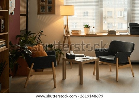 Small square table with box of tissues, paper documents and notepad of psychologist standing between two armchairs in office