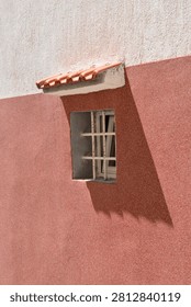 A small square window with metal bars and a tiled roof canopy built into a textured two-tone plaster house wall casting a geometric shadow