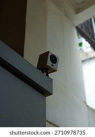 A small, square-shaped security camera (CCTV) mounted high on an exterior corner or ledge, observing an entrance or open courtyard with bright daylight.