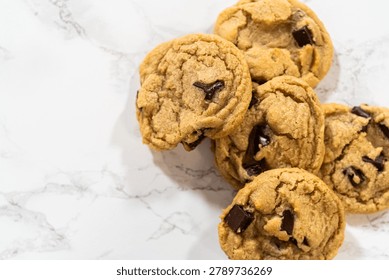 A small stack of chocolate chip cookies sits on a white marble surface. Overhead lighting highlights texture and baked finish.