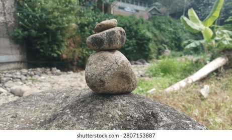 A small stack of smooth river stones carefully balanced on a large rock in a peaceful natural setting. The background features lush green vegetation, a dry rocky streambed, and a few houses partially visible among the trees. The image symbolizes balance, patience, and harmony with nature, with a softly blurred background that draws attention to the stone arrangement.