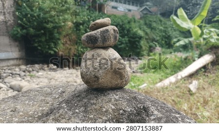A small stack of smooth river stones carefully balanced on a large rock in a peaceful natural setting. The background features lush green vegetation, a dry rocky streambed, and a few houses partially visible among the trees. The image symbolizes balance, patience, and harmony with nature, with a softly blurred background that draws attention to the stone arrangement.