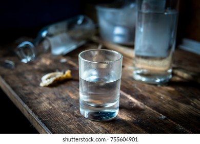 A small stack of vodka close up on the old dirty wooden table amid empty bottles lying on the table. Alcoholism concept.