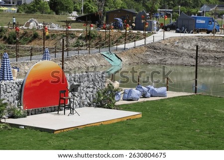 A small stage with an orange backdrop is located on the grass, surrounded by white stone walls and greenery. In front of it stands a pond. 