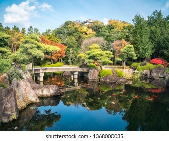 Small stone bridge over the pond full of koi fish at Koko-en Garden in autumn, with the rooftop of Himeji Castle just peeking over the tree line, in Japan.