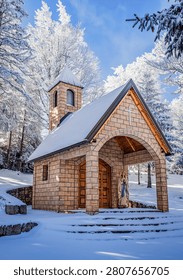 Small stone chapel surrounded by snow covered forest under a bright blue winter sky. Peaceful mountain landscape with frosted trees, Christian architecture, spirituality and seasonal nature.
