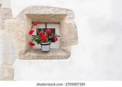 a small stone-framed window with a pot of red flowers with green leaves, on a whitewashed facade, copy space, geraniums, Pelargonium