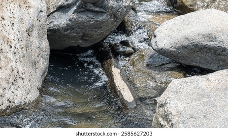 A small stream flowing between large rocks, with a piece of wood caught in the current.