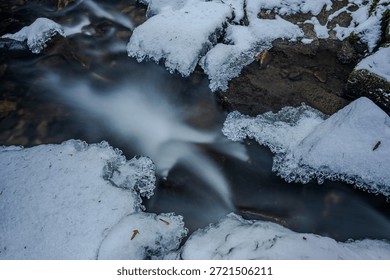 A small stream flows between ice and snow. The cold has frozen the water and the surrounding area, while it remains in motion. Small ice crystals are visible. 