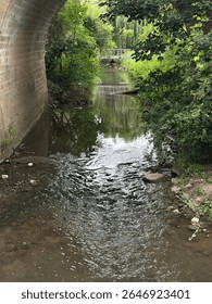 A small stream flows gently through a picturesque arched stone bridge, creating a peaceful natural scene that combines rustic architecture with the soothing beauty of flowing water