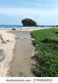 A small stream outlet flowing through a sandy beach into the ocean with a rocky islet background