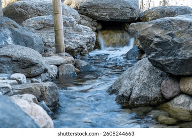 A small stream of water flows between large rocks. The rocks are grey and black. The water is clear and calm