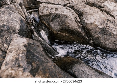 A small stream of water flows between two large rocks. The water is clear and calm, and the rocks are rough and jagged. The scene is peaceful and serene