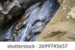 A small stream of water flows down a rocky hillside. The water is clear and calm, and the rocks are jagged and rough. The scene is peaceful and serene