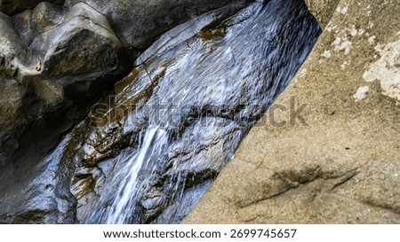 A small stream of water flows down a rocky hillside. The water is clear and calm, and the rocks are jagged and rough. The scene is peaceful and serene