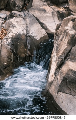A small stream of water flows down a rocky cliff. The water is clear and calm, and the rocks are jagged and rough. The scene is peaceful and serene