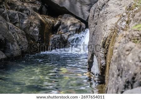 A small stream of water flows through a rocky area. The water is clear and calm, and the rocks surrounding it are large and jagged. The scene is peaceful and serene