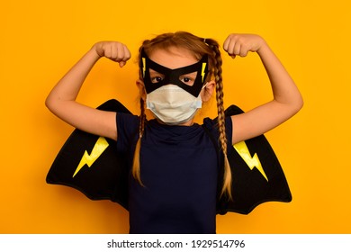 A Small Strong Pretty Girl In A Superhero Carnival Costume In A Black Mask And Wings And In A Medical Mask Clenches Her Hands Into Fists To Show Her Strength. The Child Fights Diseases And Infections