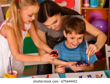 Small Students With Teacher Finger Painting In Art School Class. Teacher Cheers Children In Kindergarten. Mom And Kids At Home. Boy First Time Getting Their Hands Dirty With Paint.