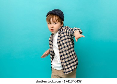 Small stylish boy, a 4-5-year-old kid in a checked shirt and a white t-shirt with a cap on his head dances with his hands spread to the side, standing out against the pastel blue background of a child