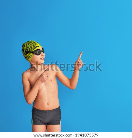 small swimmer wearing a swimming cap, glasses and a bathing suit on a blue background. child points his finger to the side, advertising. boy is studying at sports school, learning to swim. Copy space