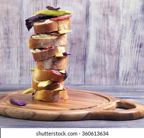 Small Tapas Sandwiches With Bread, Cheese, Sausage Meat, Avocado And Basil On A Light Wooden Cutting Board Round Table Stacked Pile In One Large High