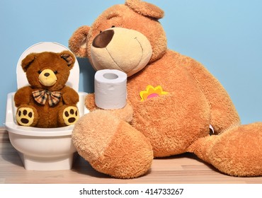 Small Teddy Bear Sitting On The Potty And Playing With Toilet Paper, Father Bear Teaching His Cute Kid How To Pee And Poo