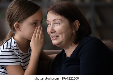 Small Teenage Girl Child Whisper In Grandmother Ear Share Secret With Grandparent At Home. Cute Little Teen Granddaughter And Elderly Granny Have Fun Talking Gossiping Together. Bonding Concept.